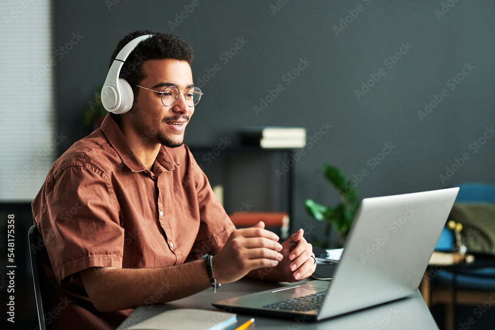 Young confident student in headphones communicating with online audience or teacher while sitting by desk in front of laptop - obrazy, fototapety, plakaty 