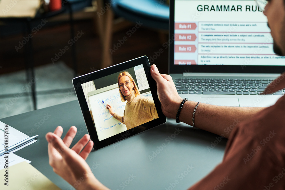 Confident blond tutor presenting new English grammar subject at lesson to online audience on tablet screen while pointing at whiteboard