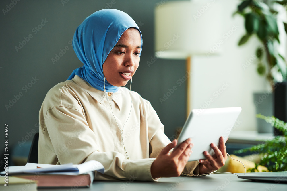 Confident Muslim female teacher or student in blue hijab looking at online audience on tablet screen while communicating to them Confident Muslim female teacher or student in blue hijab looking at online audience on tablet screen while communicating to them