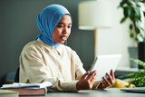 Confident Muslim female teacher or student in blue hijab looking at online audience on tablet screen while communicating to them