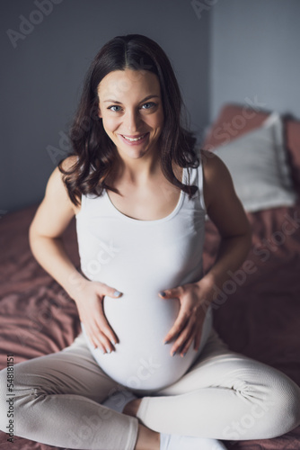 Happy pregnant woman relaxing at home. She is sitting on bed in bedroom.