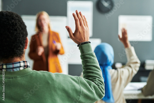 Part of back of young male student in green pullover raising hand at lesson to ask question to teacher or give his answer photo