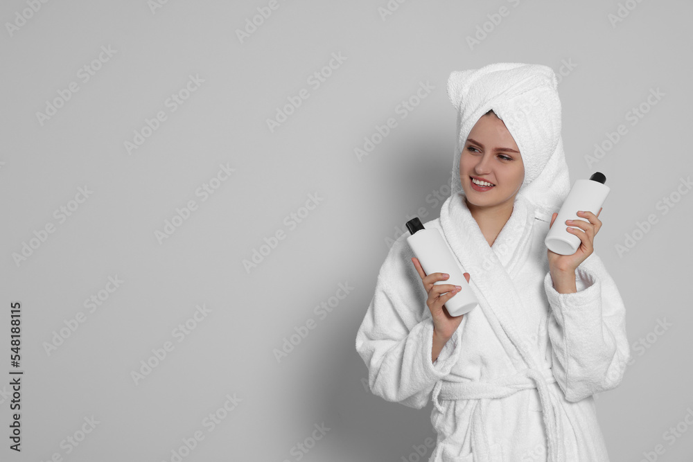 Beautiful young woman in bathrobe holding bottles of shampoo on light grey background. Space for text Beautiful young woman in bathrobe holding bottles of shampoo on light grey background. Space for text