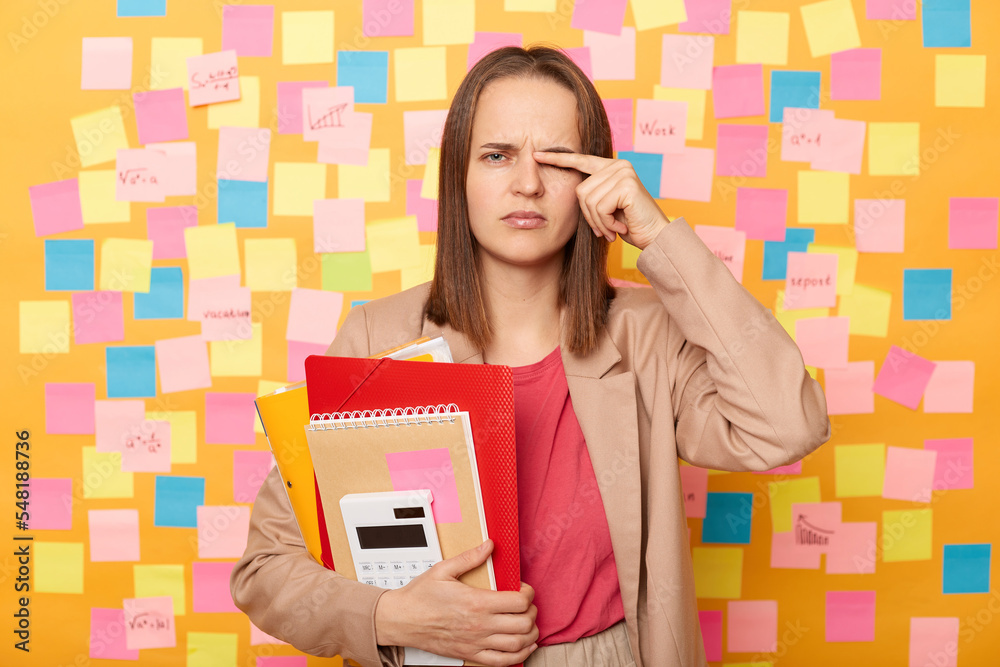 Portrait of tired upset young adult woman holding paper documents and calculator, posing against yellow wall with colorful memo cards, feels pain in eyes, working hard long hours. Portrait of tired upset young adult woman holding paper documents and calculator, posing against yellow wall with colorful memo cards, feels pain in eyes, working hard long hours.