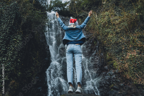 Young woman in a Santa Claus hat stands on a stone near a waterfall, raises her hands up and feels freedom, happiness. Concept of travel and wanderlust, celebration. Copy space. Mirveti Waterfall