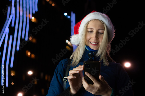 Young happy woman in a Santa Claus hat reads sms on the phone on Christmas Day on a lighted street at night in the city