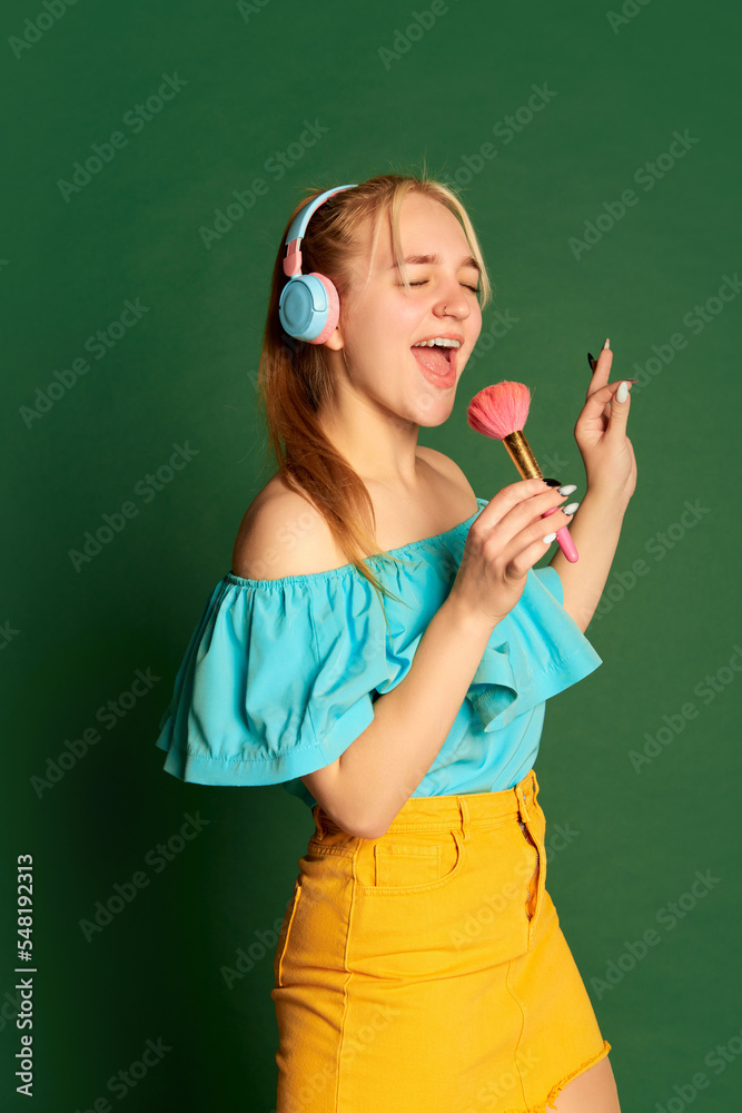 Funny singer. Half-length portrait of young beautiful girl, student in summer outfit having fun isolated over green background. Positive emotions, happiness, hobbies and joy Funny singer. Half-length portrait of young beautiful girl, student in summer outfit having fun isolated over green background. Positive emotions, happiness, hobbies and joy