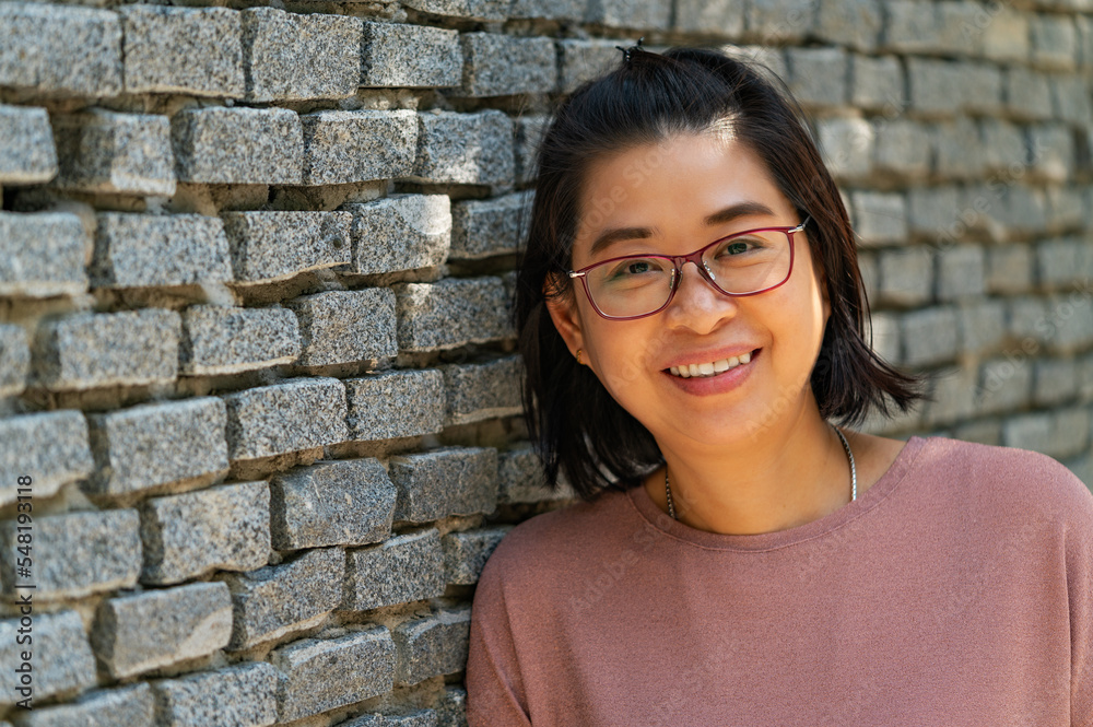 Portrait Asian middle-aged woman with smiling face, smiling, looks healthy, wears eyeglasses, stand lean on grey brick wall, perspective view, empty space for copy.