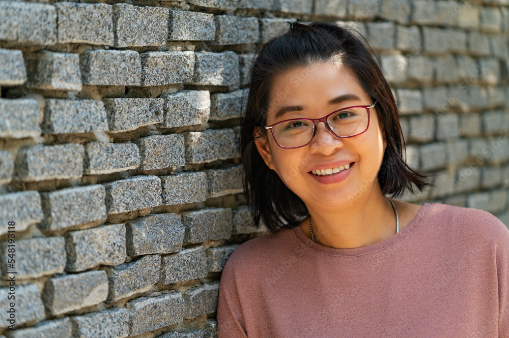 Portrait Asian middle-aged woman with smiling face, smiling, looks healthy, wears eyeglasses, stand lean on grey brick wall, perspective view, empty space for copy. Portrait Asian middle-aged woman with smiling face, smiling, looks healthy, wears eyeglasses, stand lean on grey brick wall, perspective view, empty space for copy.