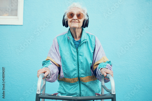 City, fashion and senior woman with headphones listening to music, audio and radio on blue background wall. Freedom, style and elderly female with modern, urban and trendy clothes with walker in town photo