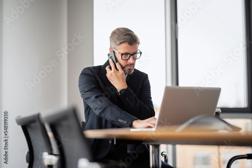 Executive middle aged businessman having a call and using laptop while working at the office. Wearing suit and tie