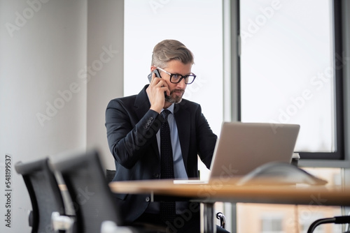 Mid adult business professional sitting at table with laptop and talking on mobile phone. Businessman using laptop while working in the office. Wearing suit and tie.