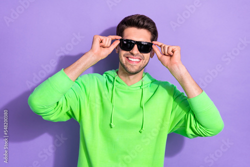 Photo of positive handsome optimistic guy brunet hairdo dressed green hoodie hands touch sunglass isolated on purple color background