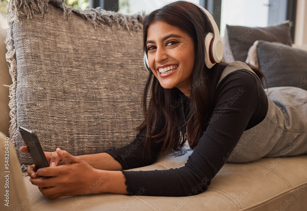 Woman, happy and headphone phone music of a person listening to a podcast or video watching at home. Portrait of an indian person with happiness and smile about mobile technology on living room sofa - obrazy, fototapety, plakaty 