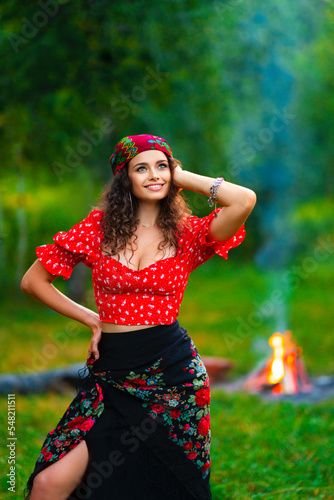 Pretty brunette curly girl in red gipsy costume and accessories posing on nature with green background and bonfire. 