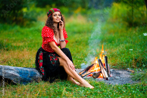 Fotografie Pretty brunette curly girl in red gipsy costume and accessories posing on nature with green background and bonfire