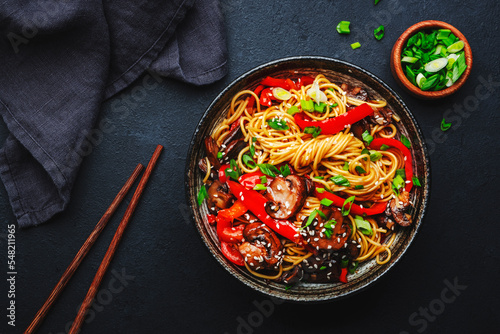 Vegan stir fry egg noodles with vegetables, paprika, mushrooms, chives and sesame seeds in bowl. Asian cuisine dish. Black table background, top view