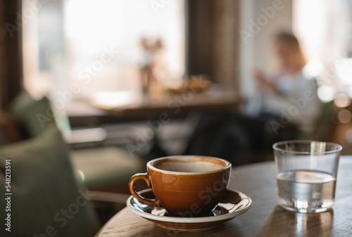 Cup and glass of water on table and blurred unrecognizable visitor having lunch on background. Cafe or restaurant concept. Copy space.