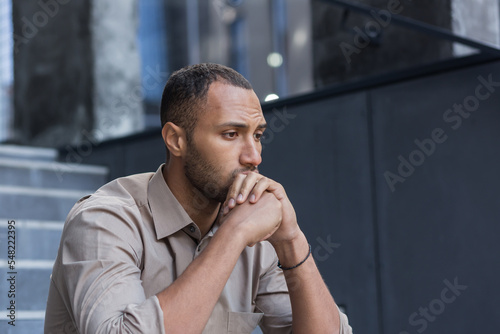 Portrait of a serious and sad hispanic.african american man. He sits sullenly outside on the stairs, holds his head in his hands, looks to the side. photo
