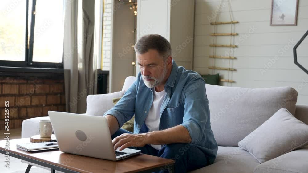 Senior old mature man typing on laptop browsing internet using apps sit on sofa alone Senior old mature man typing on laptop browsing internet using apps sit on sofa alone