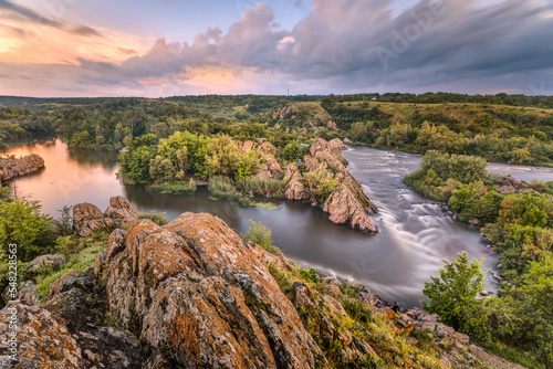 Beautiful summer sunset at the river with orange sky, clouds, rocks, green trees, flowers and water flow with reflection. Ukraine nature water background