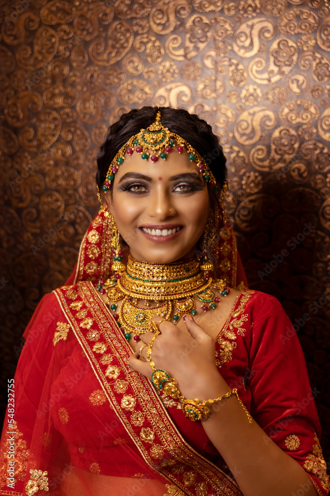 Beautiful Indian bride dressed in Hindu traditional wedding clothes Red lehenga embroidered with gold and a veil tender posing outside with golden accessories. dark background photoshoot