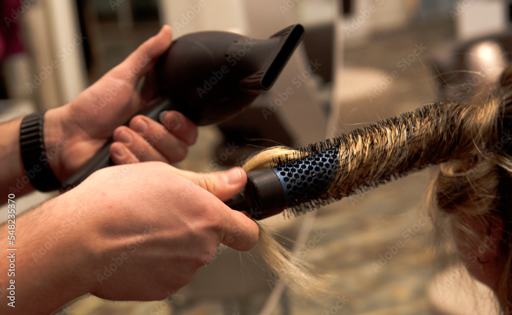 mano di parrucchiere c che con sapiente capacità arrotola una ciocca di capelli biondi a una spazzola con il phon la scalda per creare un ricciolo - obrazy, fototapety, plakaty 