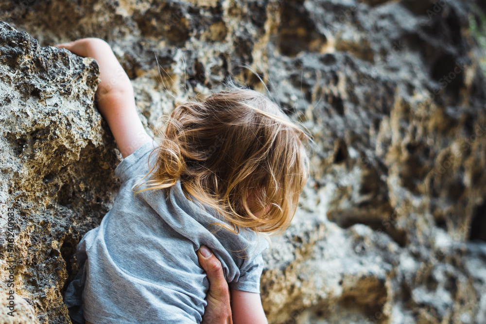 Small child rock climbing with his father supportive hand holding and helping him while on a hike in a natural park. Overcoming difficulty while developing motor skills in contact with nature.