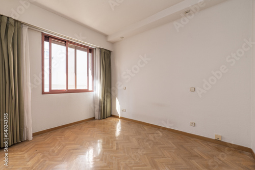 Empty living room with french oak parquet flooring with bare white walls and red aluminum window with views and white and light green sheers