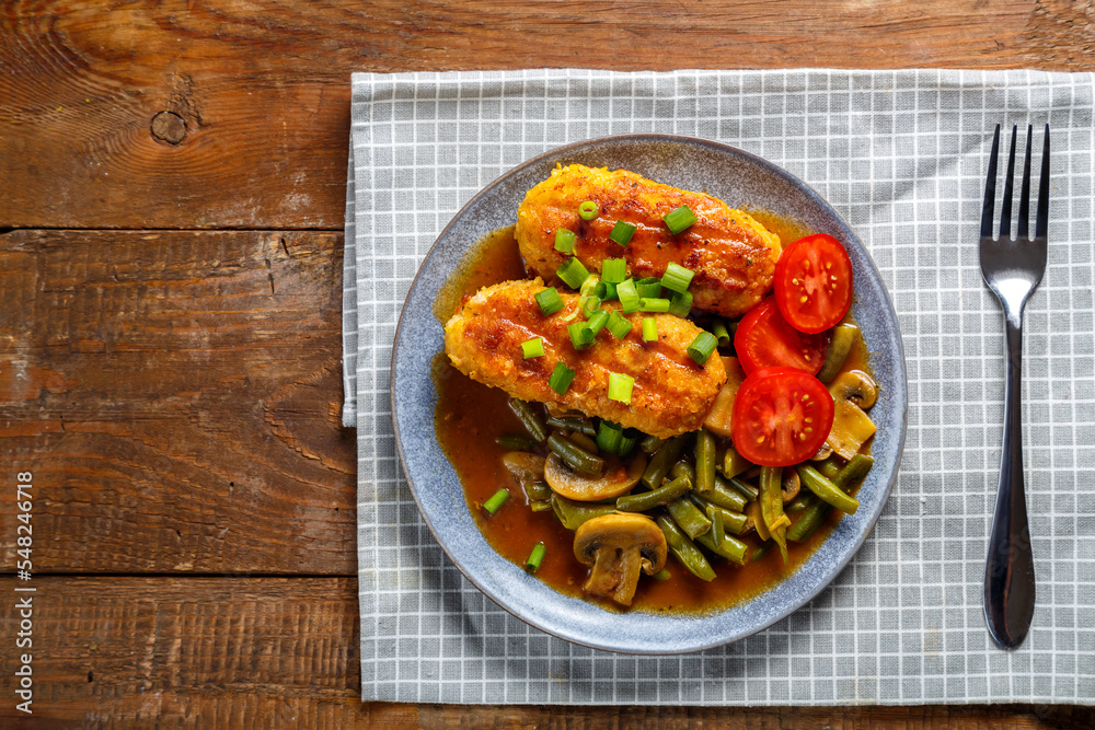 Green beans with champignons and cutlets sprinkled with green onions in a plate on a wooden table on a gray checkered napkin next to a fork. Green beans with champignons and cutlets sprinkled with green onions in a plate on a wooden table on a gray checkered napkin next to a fork.