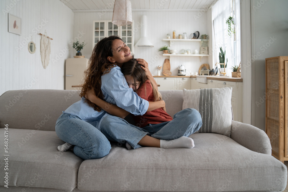 Friendly loving mother and daughter hugging sits on sofa in spacious living  room of own home. Happy smiling Caucasian woman cuddles distressed teen  girl after breakup or long-awaited reconciliation foto de Stock