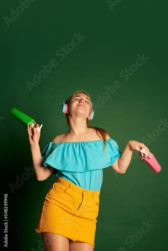 Music, dance. Portrait of young beautiful girl, student in summer outfit posing isolated over green background. Positive emotions, happiness, hobbies and joy