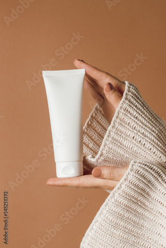 Woman hands holding white plastic tube of cream or lotion, brown background, aesthetic skincare
