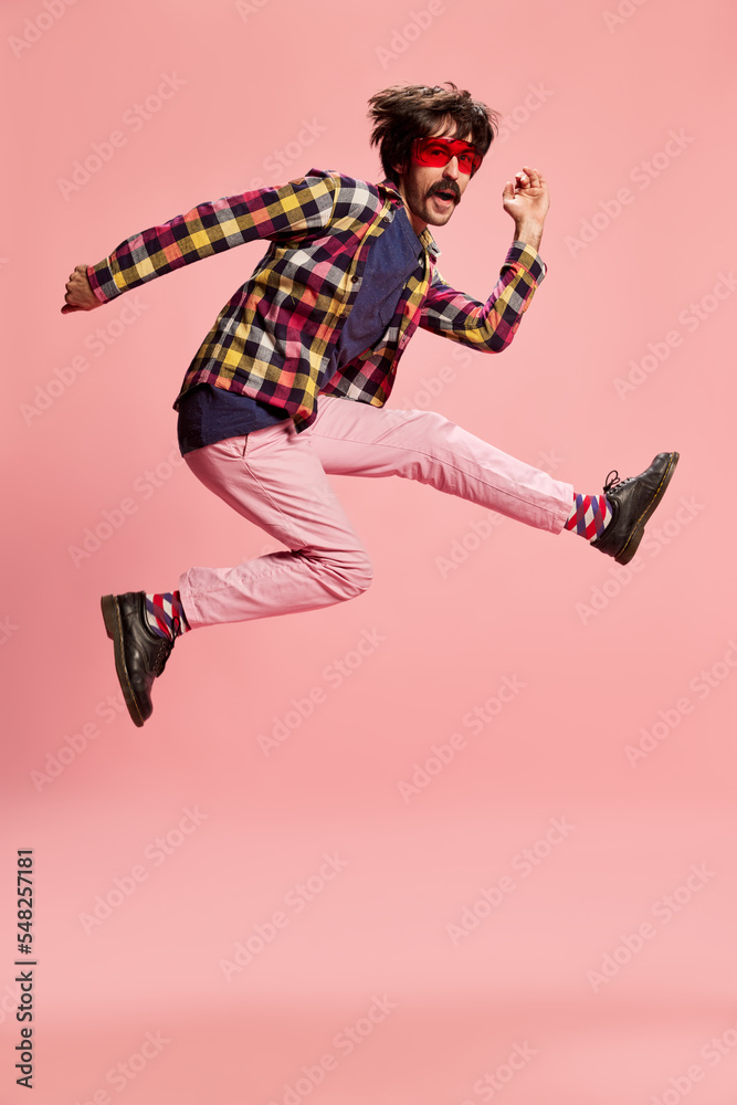 Portrait of stylish man with moustache, jumping, posing in checkered shirt and pink pants isolated over pink background