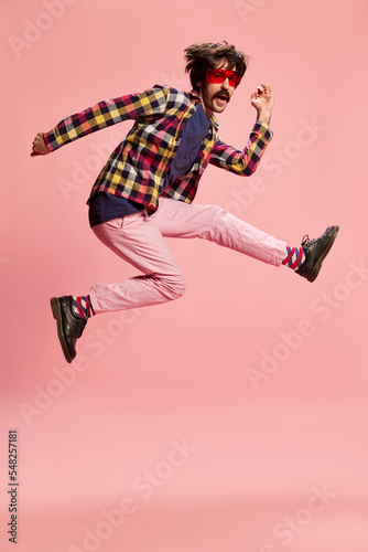 Fototapeta Naklejka Na Ścianę i Meble -  Portrait of stylish man with moustache, jumping, posing in checkered shirt and pink pants isolated over pink background