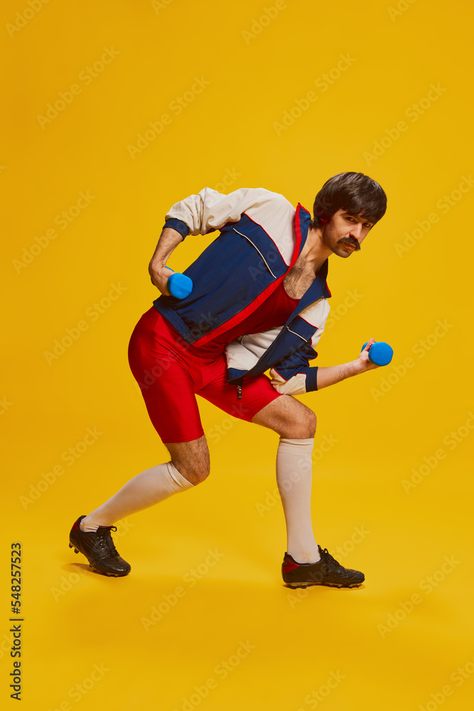 Portrait of stylish man with moustache posing in vintage sportswear training with dumbbells isolated over yellow background. Strong hands