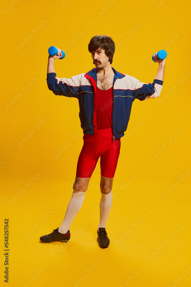 Portrait of stylish man with moustache posing in vintage sportswear training with dumbbells isolated over yellow background Portrait of stylish man with moustache posing in vintage sportswear training with dumbbells isolated over yellow background