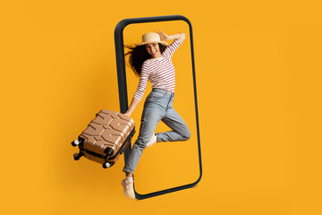 Smiling excited young arab lady tourist in hat with suitcase jumping near huge smartphone, ready for travel
