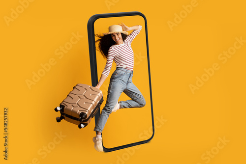 Smiling excited young arab lady tourist in hat with suitcase jumping near huge smartphone, ready for travel photo