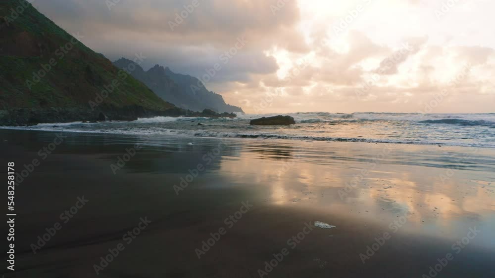 Heavenly sunset light is reflected on wet smooth sand of tropical ocean beach. Relaxing soothing aerial flight. Sunset clouds on sky behind mountain range Anaga with calm sea waves.