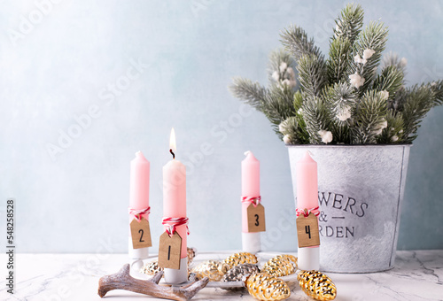 Winter holidays postcard. Advent candles. Pink burning candle, golden and silver decorative cones and bucket with branches oh fir tree against blue textured wall. Selective focus. Place for text.