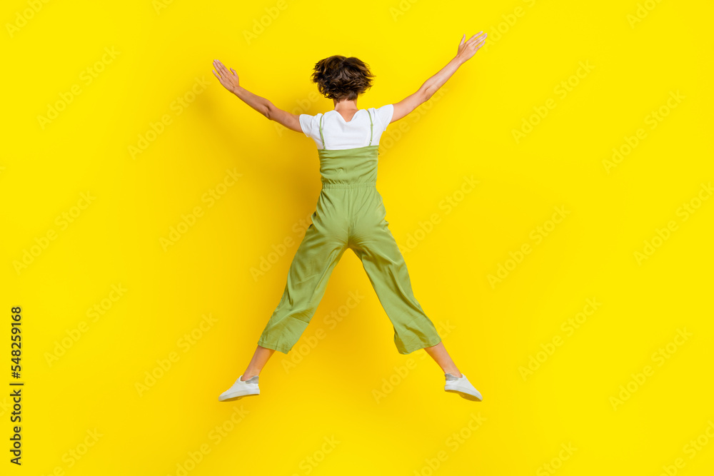 Full body size photo of funny woman dressed khaki overalls hands up star positive mood jumping high carefree isolated on yellow color background