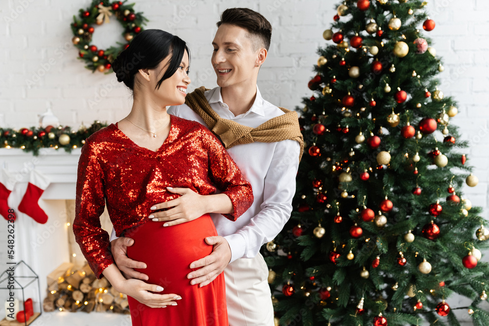 cheerful man and pregnant asian woman in elegant clothes smiling at each other in living room with Christmas decor cheerful man and pregnant asian woman in elegant clothes smiling at each other in living room with Christmas decor