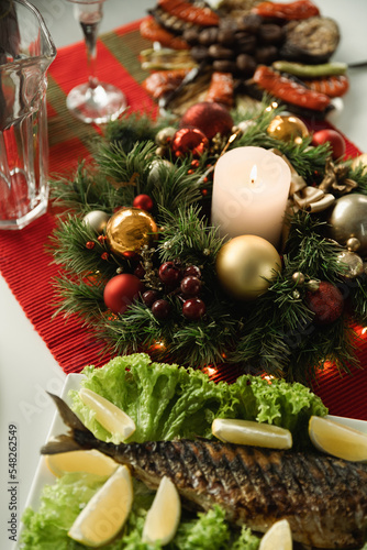 Christmas wreath with shiny baubles and lit candle near grilled fish with sliced lemon served for festive dinner