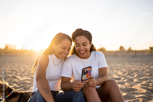 Hispanic lesbian couple use a phone and sit on a blanket at the beach with a dog photo