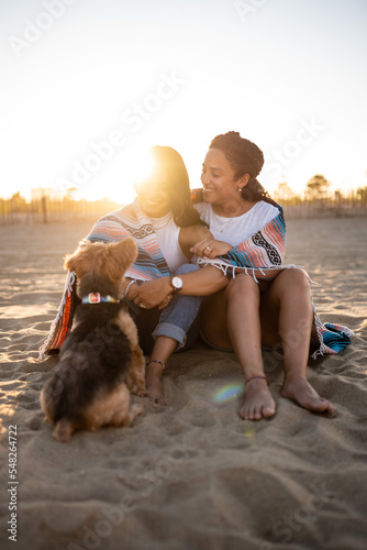 Hispanic lesbian couple sit on the beach with a blanket and dog