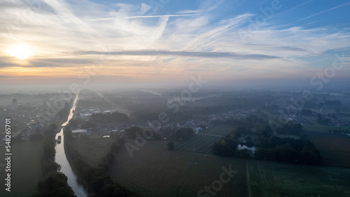 Aerial view shot by a drone of land or landscape of green fields under a misty morning sunrise sky. Include agriculture farm, house building, village. That real estate or property. Plot of land for