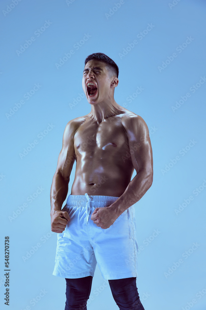 Portrait of young muscular man training, shouting to rise sportive spirit isolated over blue background in neon light