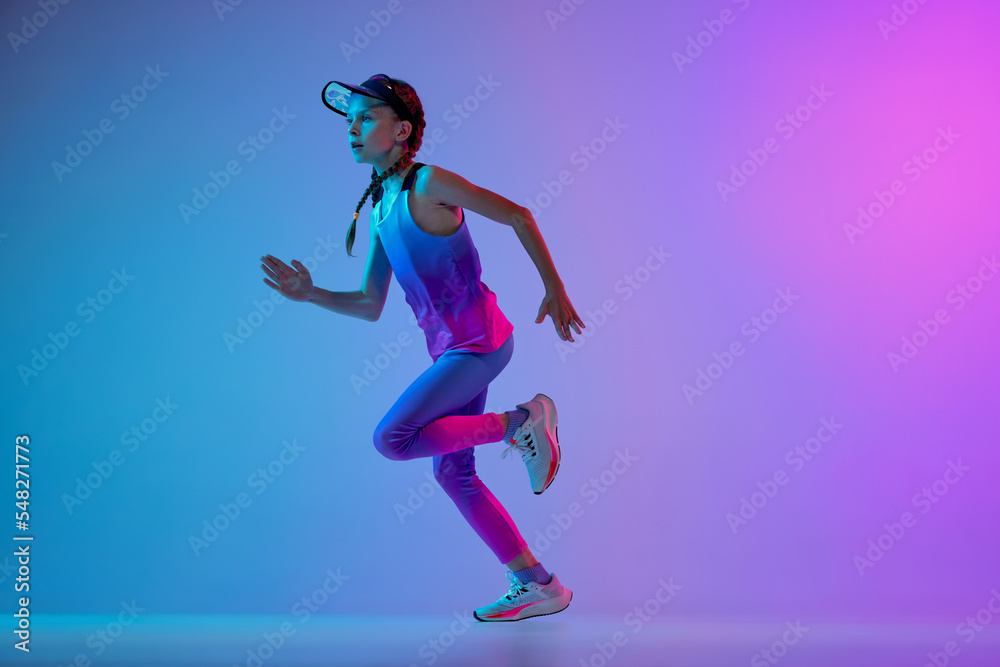 Champion. Sportive little girl, junior runner in stylish sportswear and cap running isolated on gradient pink-blue background in neon. Concept of sport, fashion, fitness and education