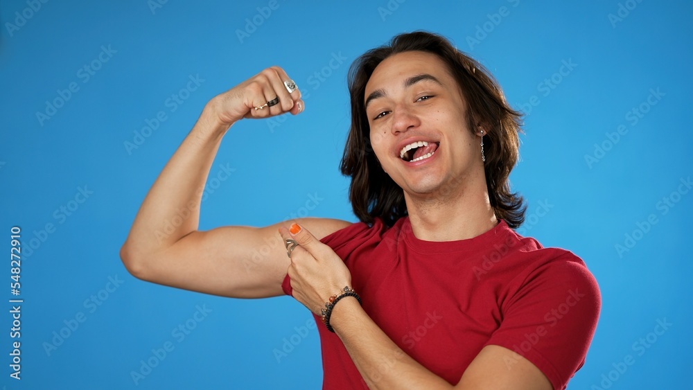 Portrait of Latino Hispanic non binary gender fluid young hipster man 20s with show muscles, have fun energy isolated on blue background in studio